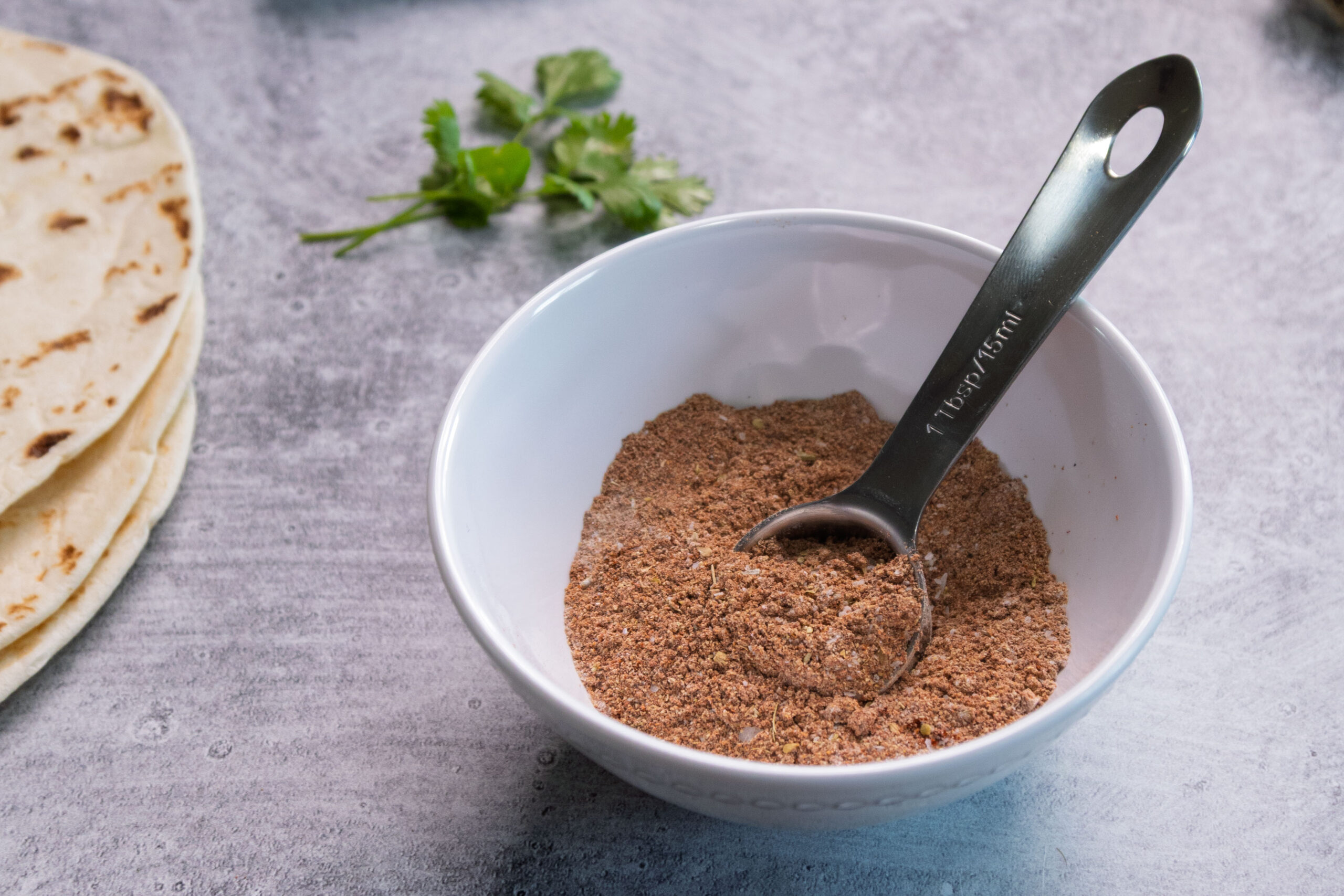 A white bowl filled with homemade taco seasoning and a metal tablespoon sits on a gray countertop, with fresh cilantro and a stack of tortillas in the background.