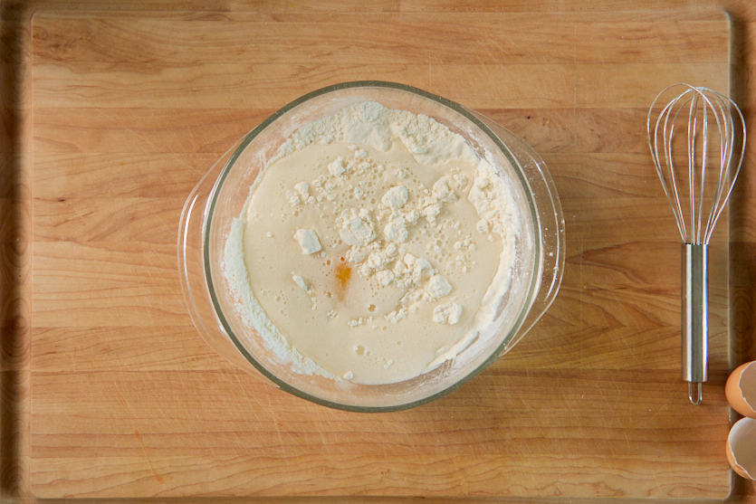 A glass bowl with flour, milk, and vanilla sits on a wooden surface—perfect for whipping up takeout classics like Tempura Chicken. Nearby are a metal whisk, cracked eggshells, and an empty egg carton. The ingredients appear ready to be mixed.
