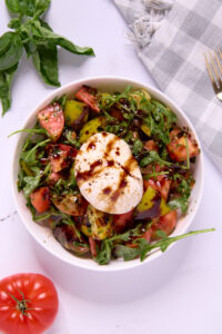 A white bowl filled with arugula, chopped tomatoes, and a whole ball of burrata cheese creates a fresh twist on Caprese Salad, drizzled with balsamic glaze. A basil sprig, tomato, and checkered napkin with a fork rest nearby on a white surface.