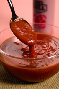 A spoon lifts thick, bbq sauce from a clear glass bowl. The sauce appears smooth and glossy. A blurred bottle is visible in the background. The bowl sits on a textured surface.