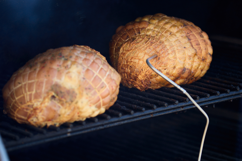 Two large, netted hams are cooking on a grill rack—one with a meat thermometer probe—perfect for those who enjoy the flavor of a pellet smoker or want to smoke turkey breast with no brine needed.