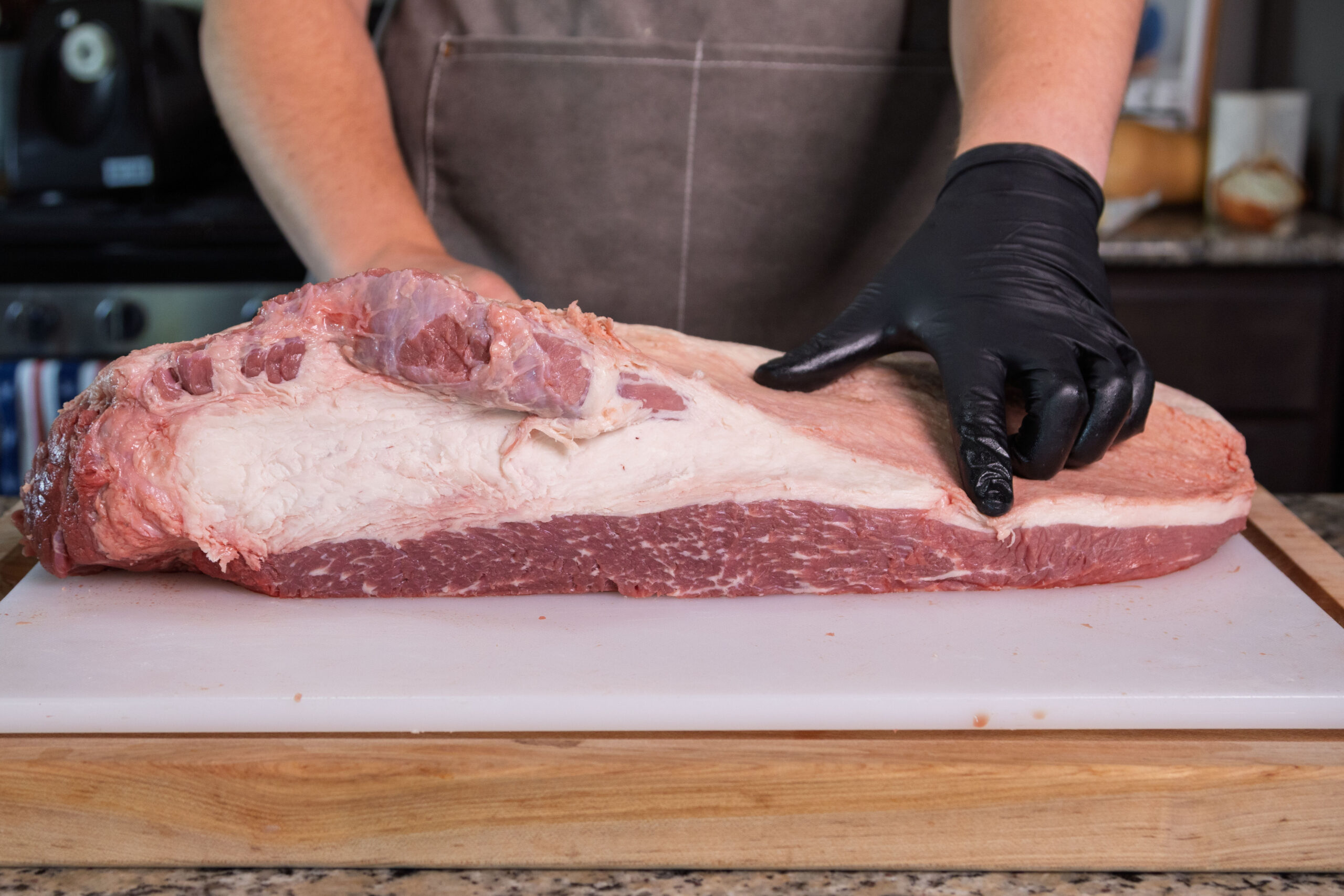 A person wearing a gray apron and black glove points at a large raw beef brisket on a white cutting board in a kitchen, ready to trim brisket as part of a step-by-step guide for preparing brisket for smoking.