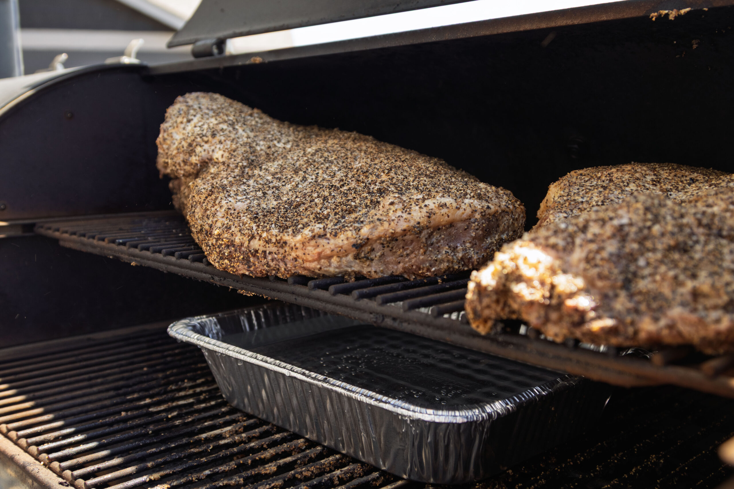 Two large pieces of brisket for smoking cook on the upper rack of a smoker grill, with a disposable aluminum tray beneath them to catch drippings.