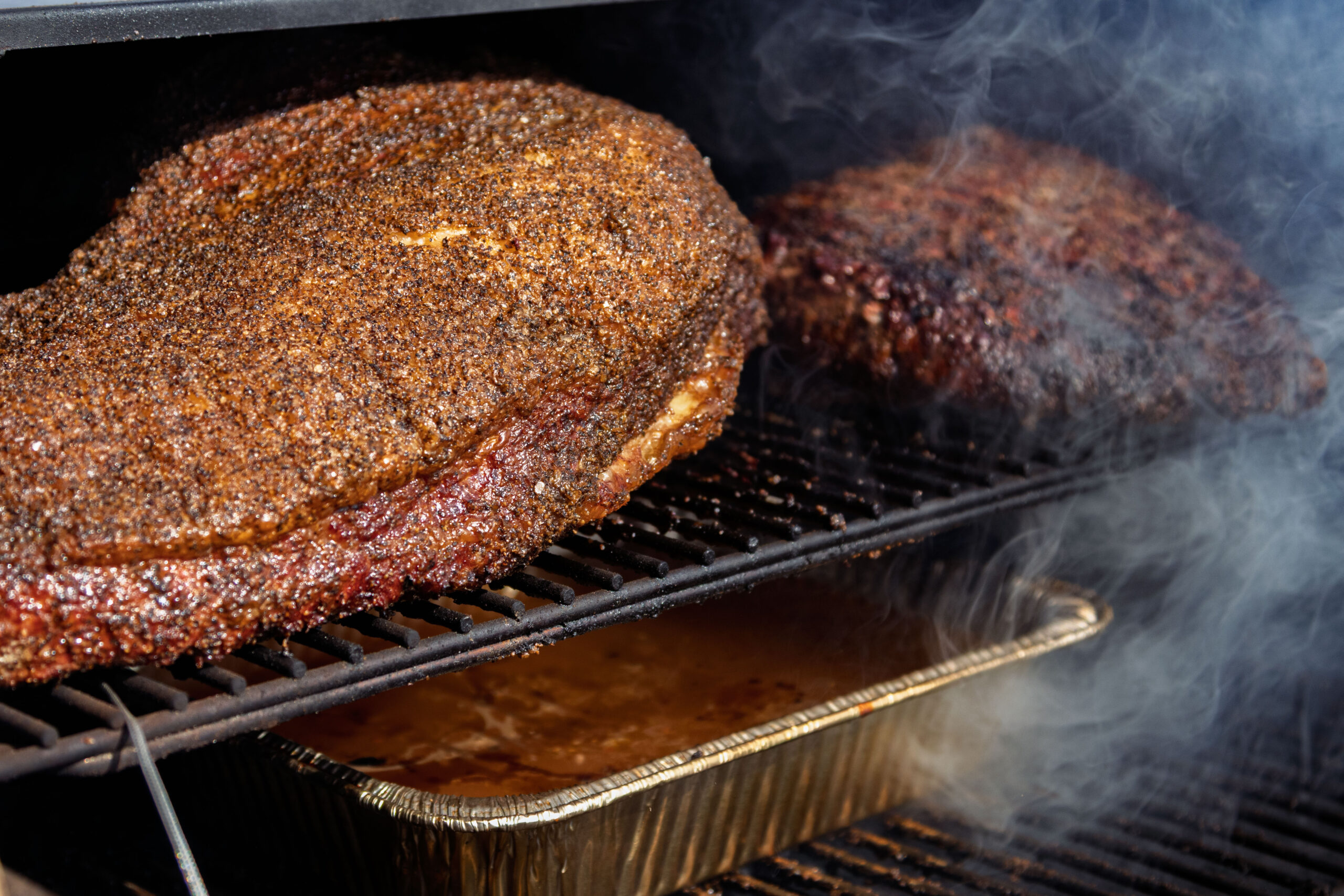 Two large cuts of seasoned meat, perfect brisket for smoking, are on a grill rack above an aluminum tray filled with liquid. Smoke surrounds the meat inside the smoker. A meat thermometer probe is inserted into one piece.