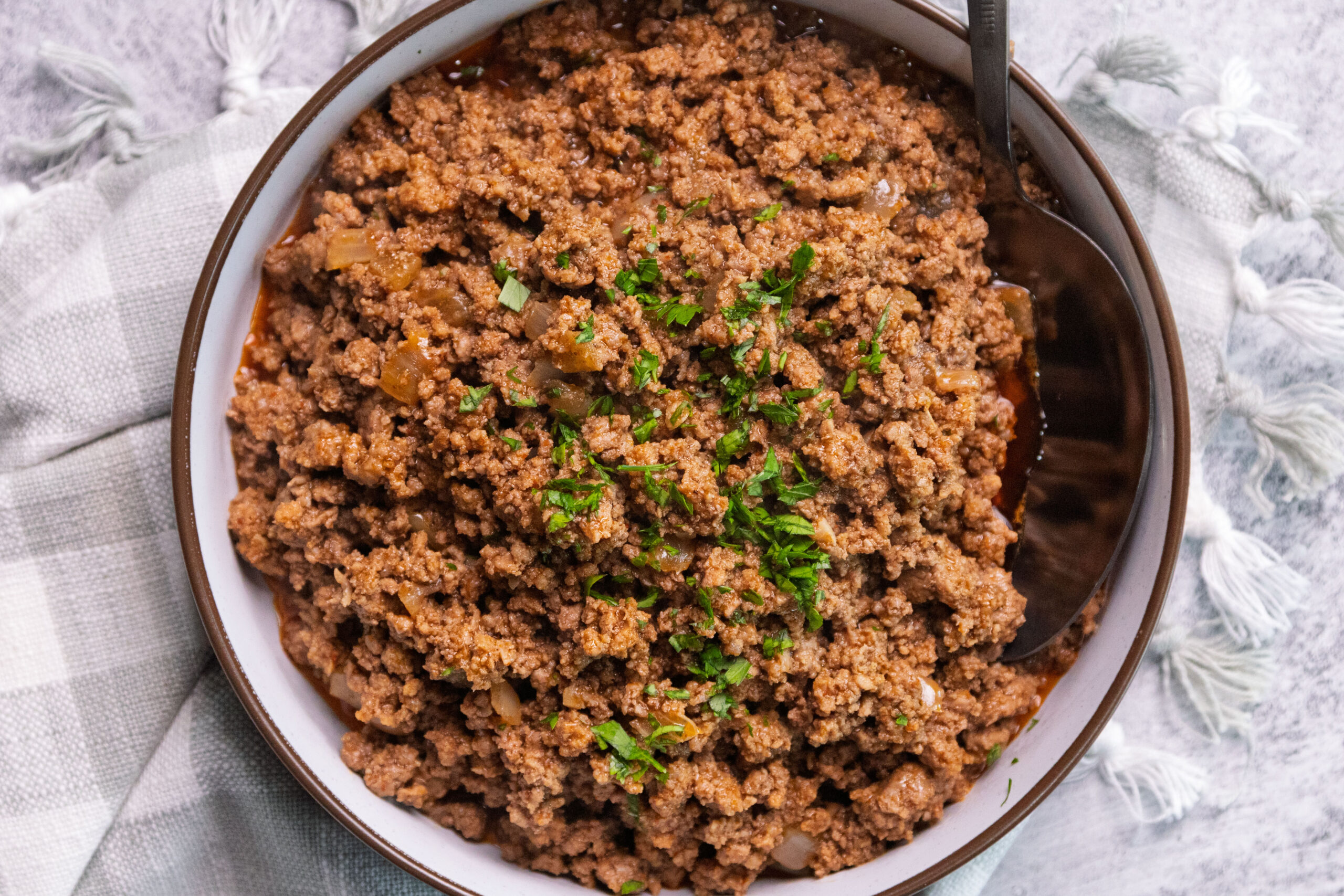 A bowl filled with flavorful ground beef taco meat mixed with small pieces of onion, topped with chopped parsley, with a spoon resting inside. The bowl sits on a gray and white checkered cloth.