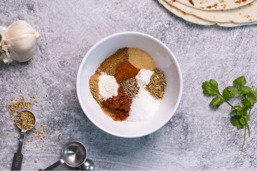 A white bowl filled with various spices including paprika, salt, black pepper, garlic powder, and dried herbs sits on a gray countertop with garlic, cilantro, measuring spoons, and tortillas nearby.