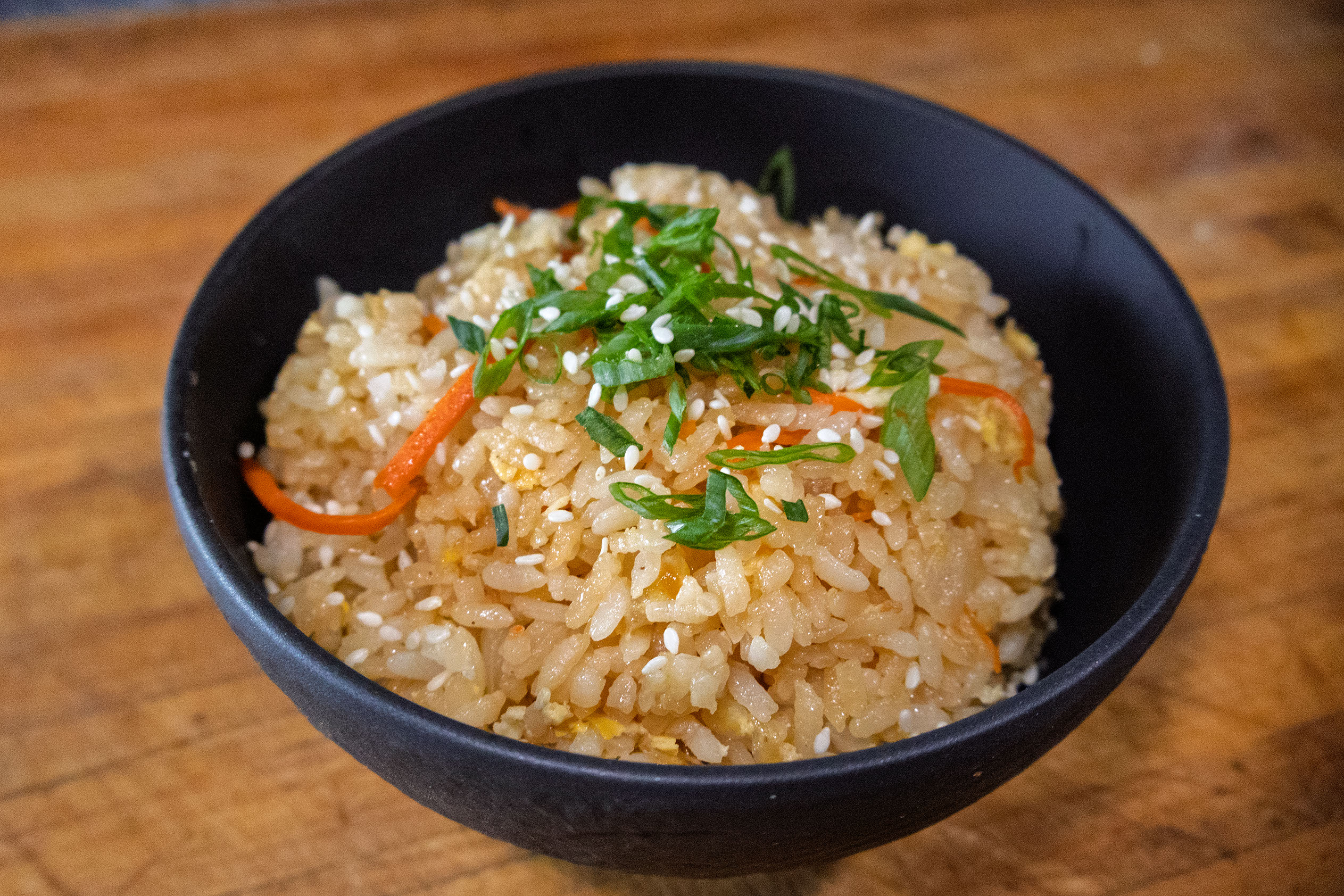 A black bowl filled with fried rice garnished with chopped green onions, shredded carrots, and sesame seeds, placed on a wooden surface.