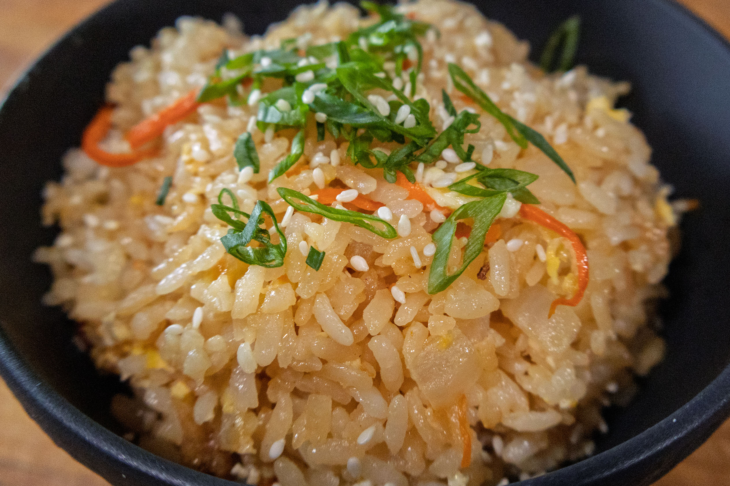 A close-up of a bowl of fried rice topped with sliced green onions, sesame seeds, and thin strips of red pepper. The rice appears golden brown and is served in a black bowl.