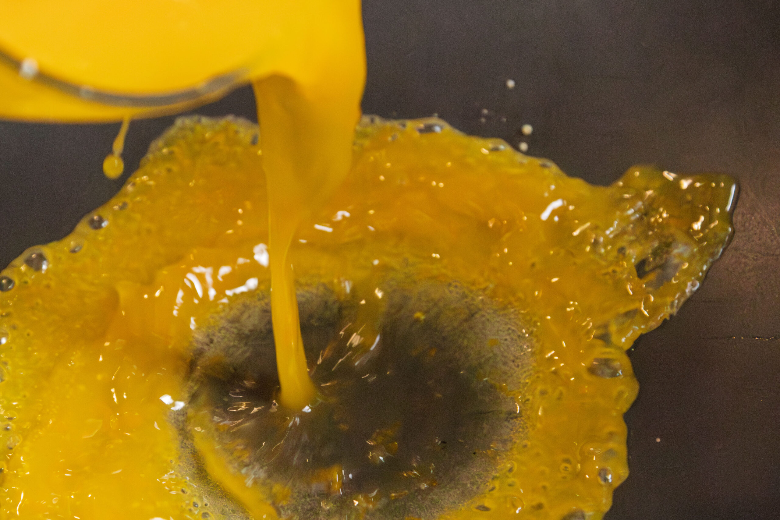 A close-up of liquid egg yolk being poured onto a dark, flat surface. The yolk splashes and spreads outward, creating an uneven, glossy yellow pool.