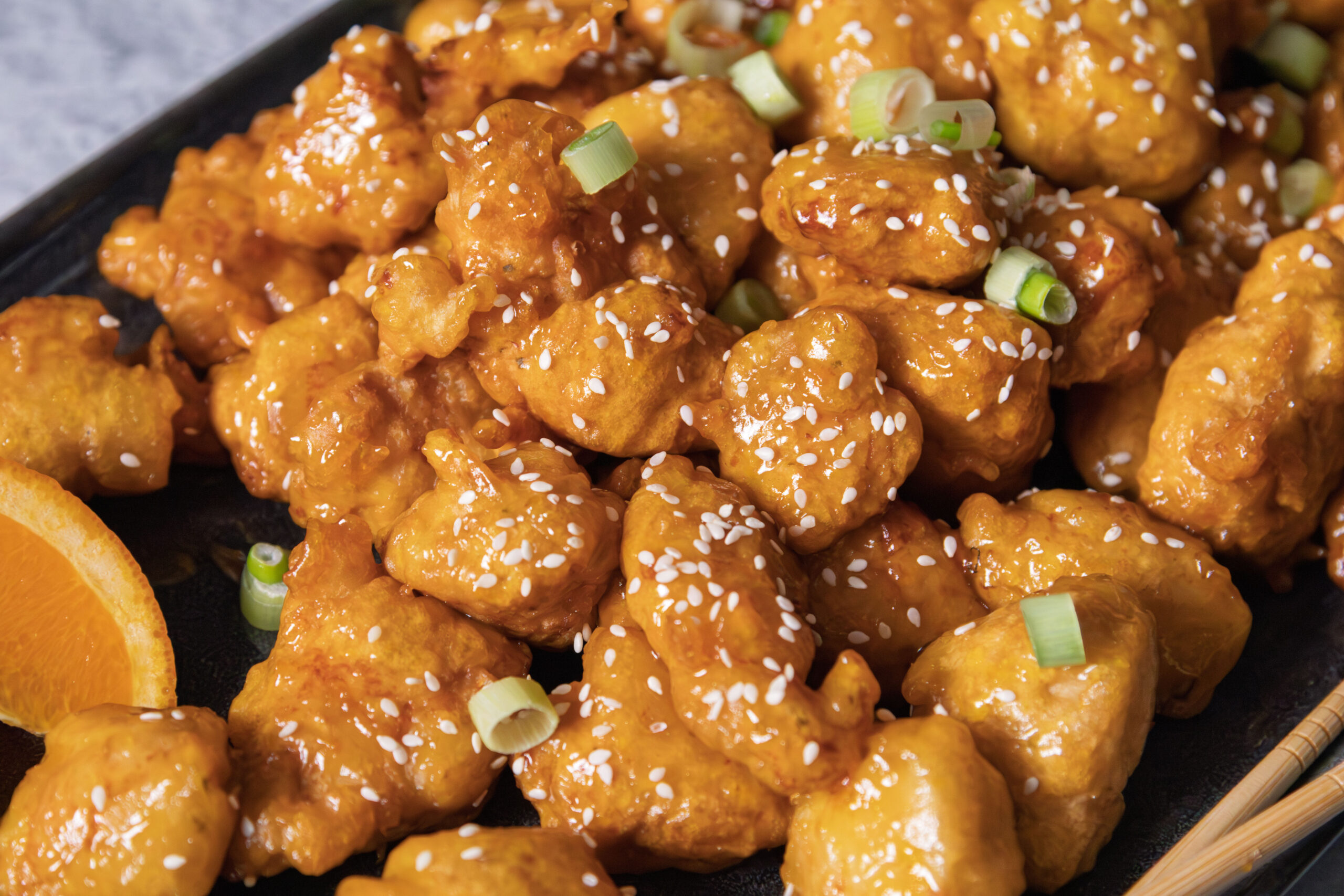 Close-up of orange chicken pieces coated in sauce, sprinkled with white sesame seeds and sliced green onions, served on a black plate with a slice of orange visible in the corner.