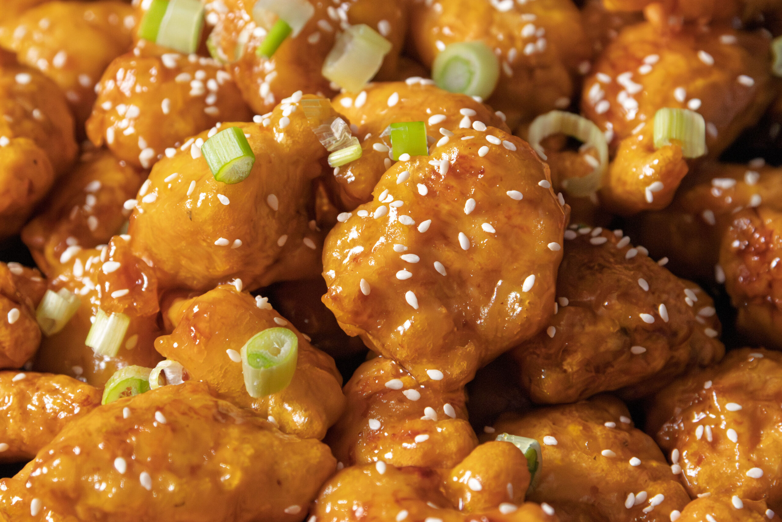 Close-up of pieces of orange chicken coated in sauce, topped with white sesame seeds and sliced green onions.