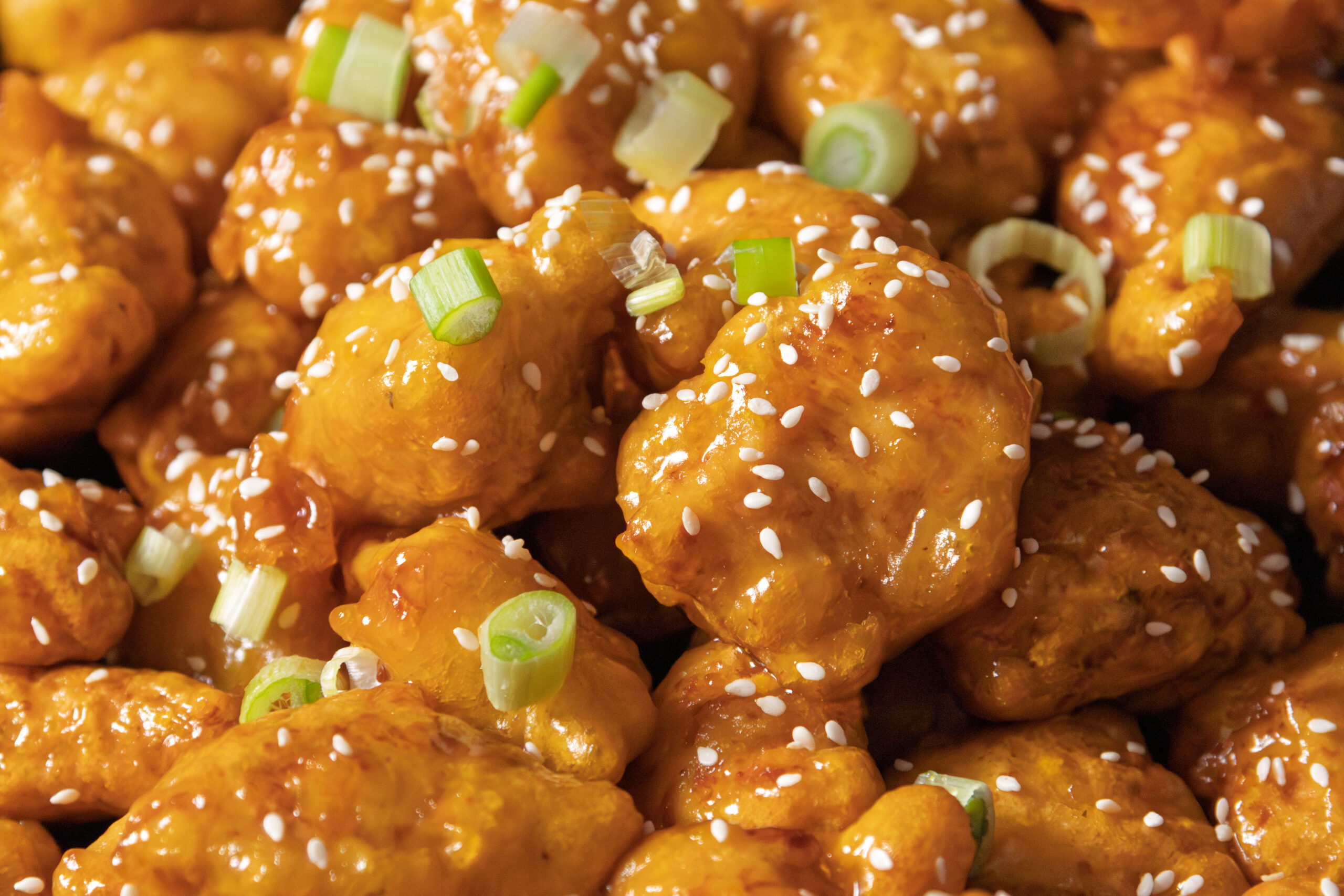 Close-up of golden, sauce-covered pieces of fried chicken topped with white sesame seeds and chopped green onions.