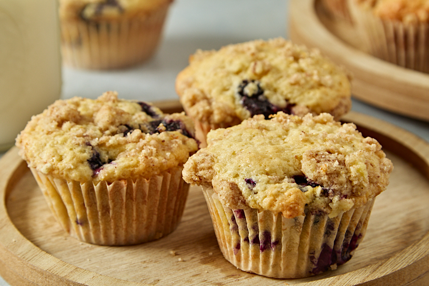 Three blueberry muffins with crumb topping are arranged on a wooden plate, with another muffin partially visible in the background. The muffins appear freshly baked and have a golden-brown color.