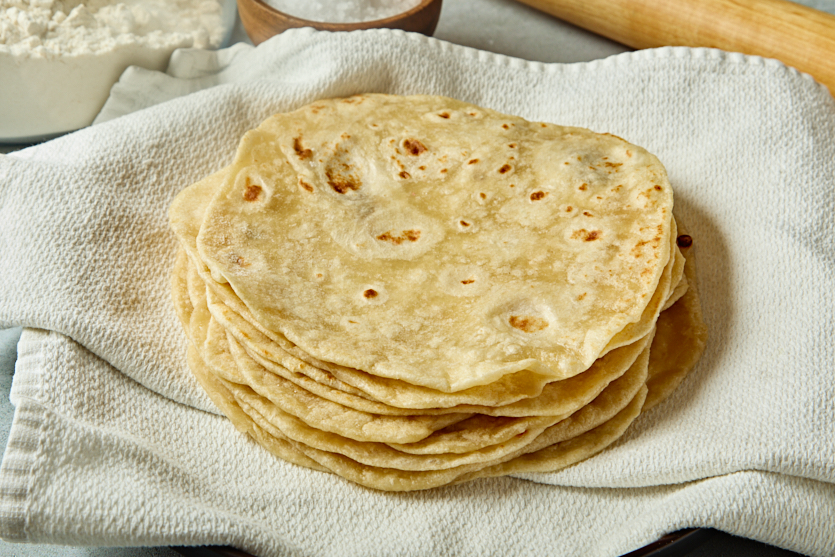 A stack of homemade flour tortillas sits on a textured white cloth, with a bowl of flour and a rolling pin visible in the background.