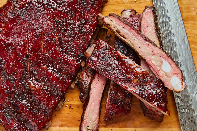 Sliced Texas-style bbq spare ribs with a dark seasoning crust are arranged on a wooden cutting board next to a large knife. The smoked spare ribs, prepared on a pellet grill, show a pink smoke ring and glistening surface.