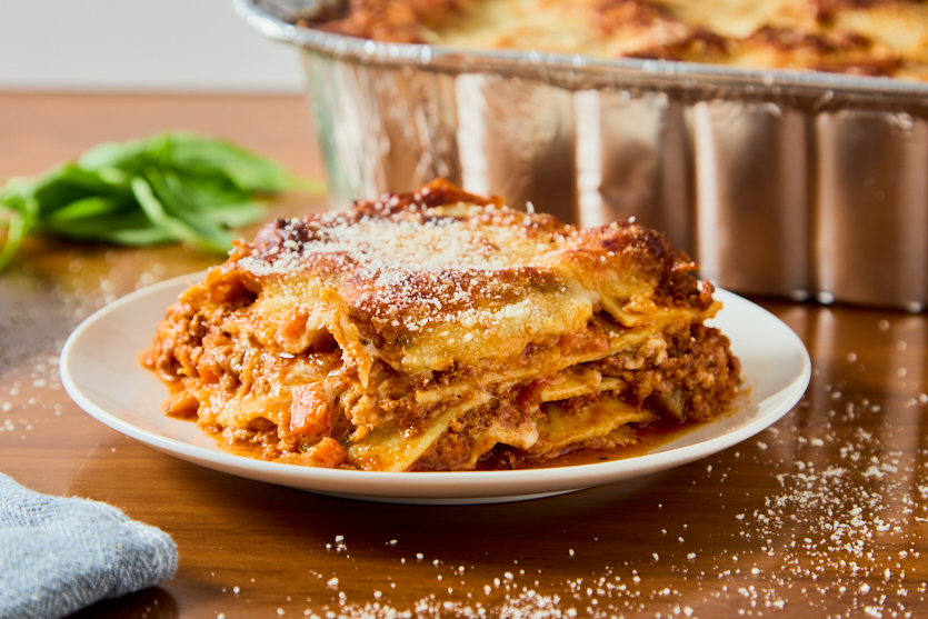 A slice of lasagna topped with grated cheese sits on a white plate, with a pan of lasagna and fresh basil in the background on a wooden table.