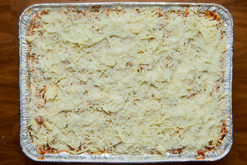A rectangular foil tray sits on a wooden surface, filled with an unbaked casserole topped with shredded cheese and breadcrumbs. The dish is ready to be cooked.