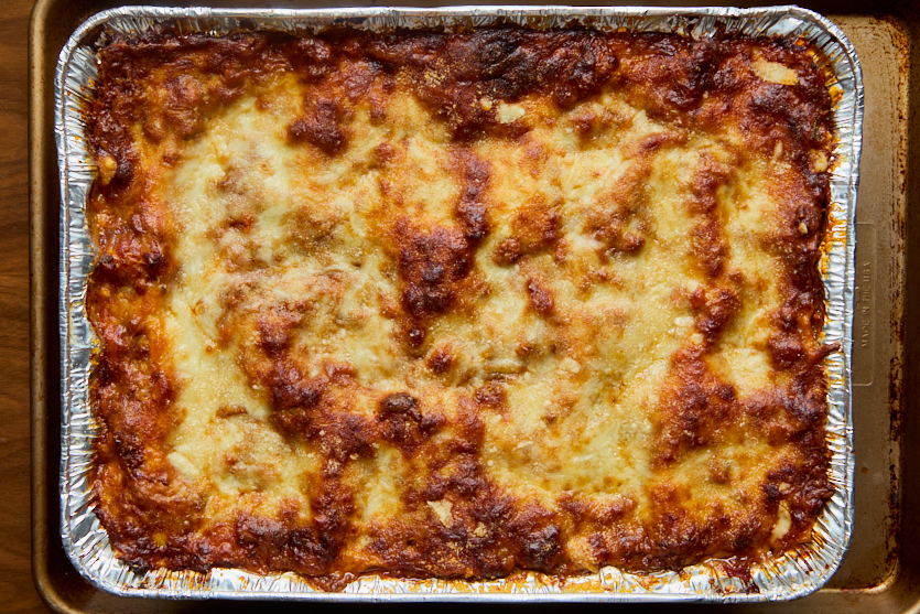 A large rectangular tray of baked lasagna sits in a foil container, with a browned, bubbly cheese layer on top and visible edges of sauce and pasta.