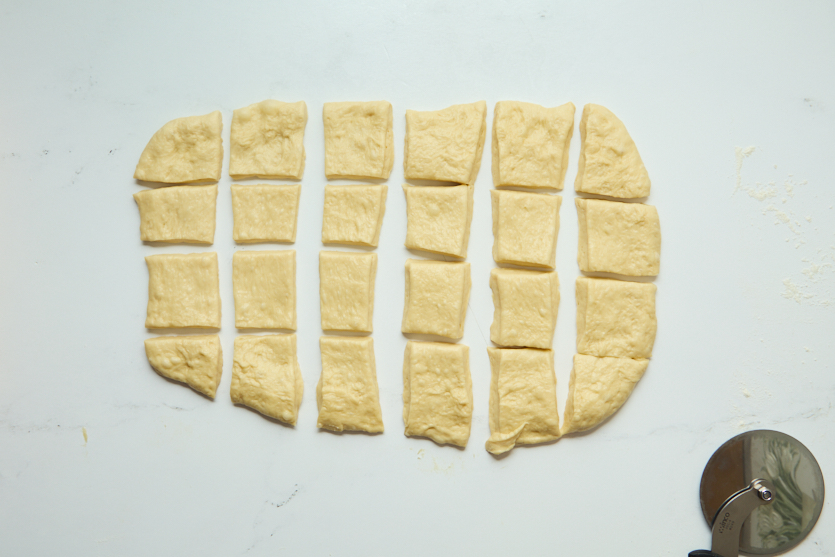 Rectangular pieces of dough, reminiscent of chewy beignets, are arranged on a white surface in a grid pattern. A metal pizza cutter is partially visible in the lower right corner.