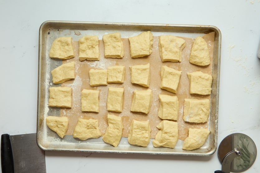A baking tray holds 24 pieces of unbaked, roughly rectangular dough spaced evenly apart—soon to become chewy beignets reminiscent of classic New Orleans beignets. A pizza cutter and a dough scraper are visible on the left and bottom edges of the tray on a white surface.