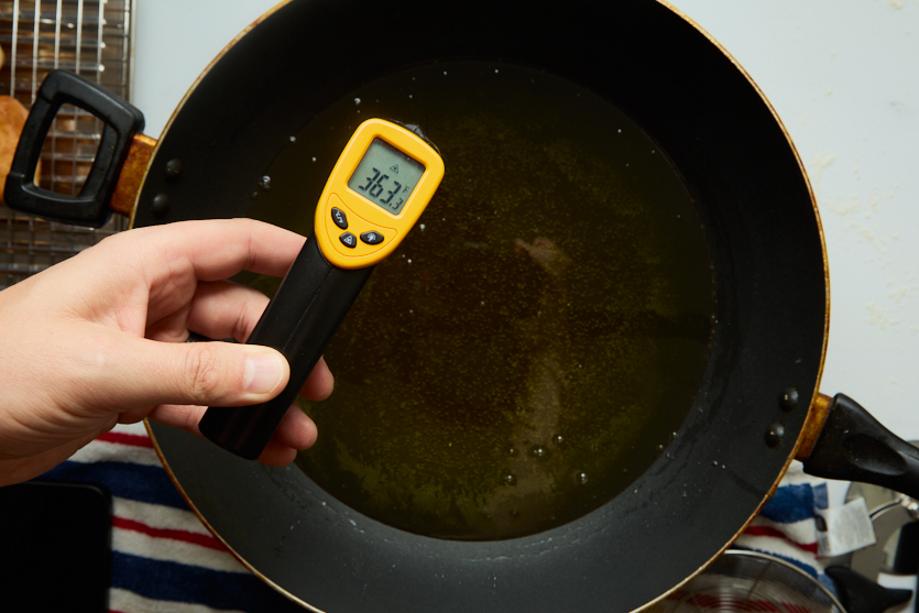 A hand holds an infrared thermometer over a pan of hot oil, displaying a temperature of 363.1 degrees Fahrenheit—the perfect heat for frying crispy beignets. A cooling rack and striped towel are visible nearby, ready to catch the freshly made New Orleans beignets as they come out of the oil.