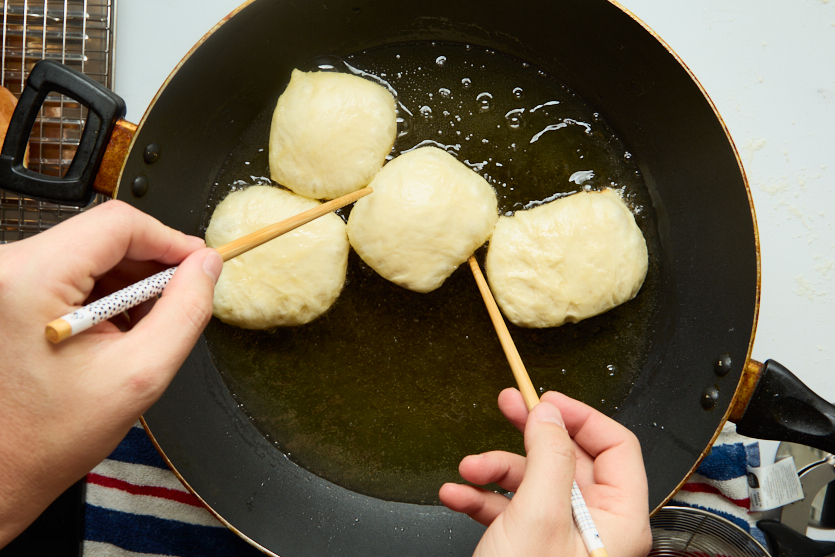 Four pieces of dough, soon to become crispy beignets, are being fried in oil in a black pan. Two hands are using chopsticks to turn or move the dough. A cooling rack sits nearby, and a striped towel is partially visible under the pan.