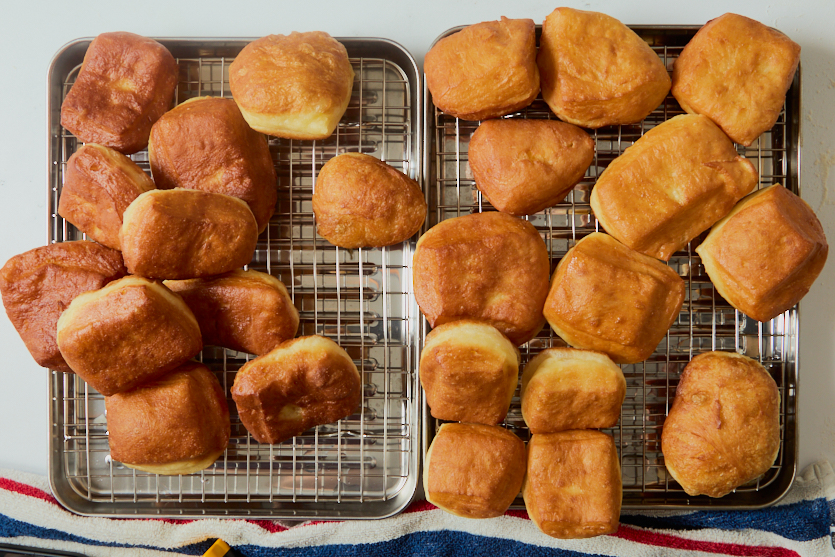 Two metal cooling racks hold golden brown, square and rectangular crispy beignets, arranged on a white surface with a striped red, white, and blue towel partially visible at the bottom of the image.
