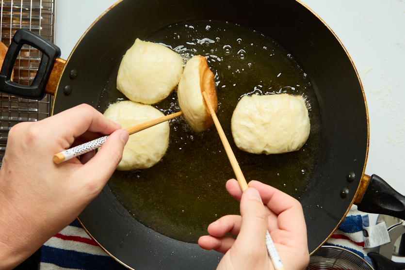Three pieces of dough are being fried in a pan of oil, transforming into crispy beignets. Two hands are using chopsticks to turn one piece of dough, revealing its golden-brown cooked side. A cooling rack and a striped towel are visible nearby, ready to hold the freshly made New Orleans beignets as they come out of the hot oil.