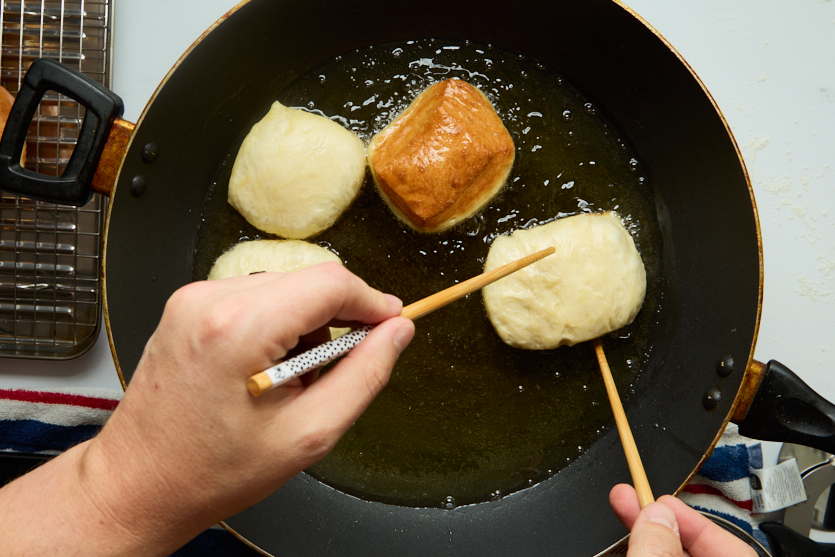 Four dough balls, soon to become crispy beignets, are being fried in hot oil in a black pan. A person uses chopsticks to turn one of the chewy beignets. A cooling rack and striped cloth are visible next to the pan.