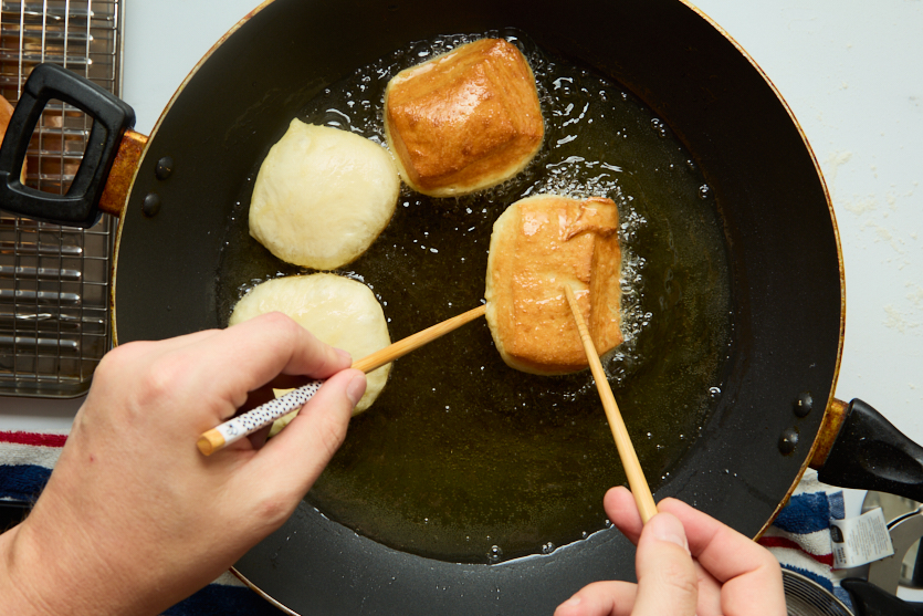 Two hands use chopsticks to fry dough pieces in hot oil in a black pan. Two pieces are golden brown, and two are still pale. A cooling rack sits beside the pan. The scene suggests the process of making New Orleans-Style Beignets, capturing that signature moment when Crispy Beignets first emerge from the oil and are set aside to cool.