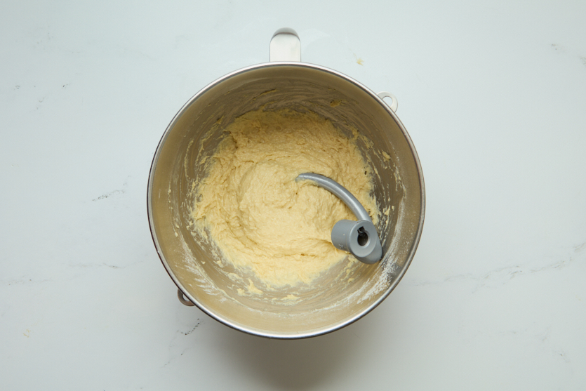 A metal mixing bowl with a paddle attachment containing a thick, pale batter for chewy beignets sits on a white countertop, viewed from above.