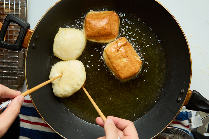 Two hands use chopsticks to fry four dough pieces in a pan of oil; two pieces are golden brown and crispy like classic New Orleans beignets, while two remain pale. A cooling rack and striped towel are nearby.