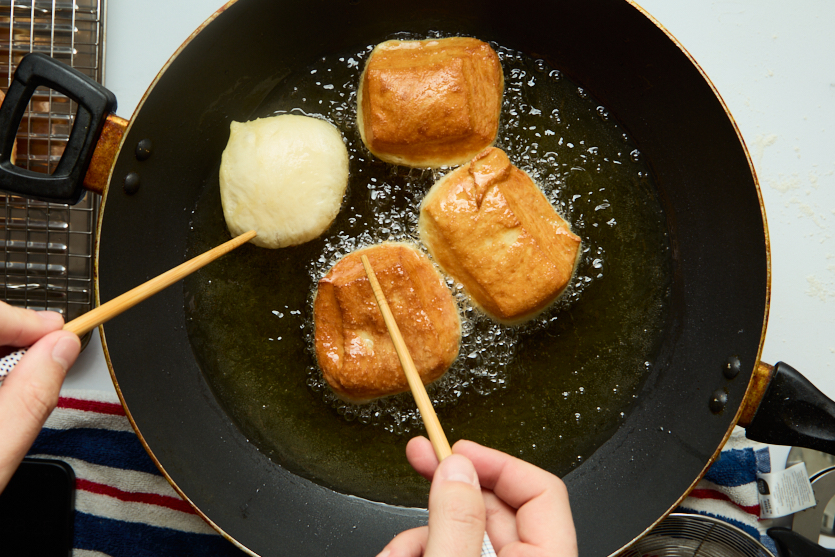 Three golden brown dough pieces and one uncooked dough piece are frying in hot oil in a pan, transforming into chewy beignets. Two hands are turning the beignets using chopsticks. A cooling rack and a striped towel are visible nearby.