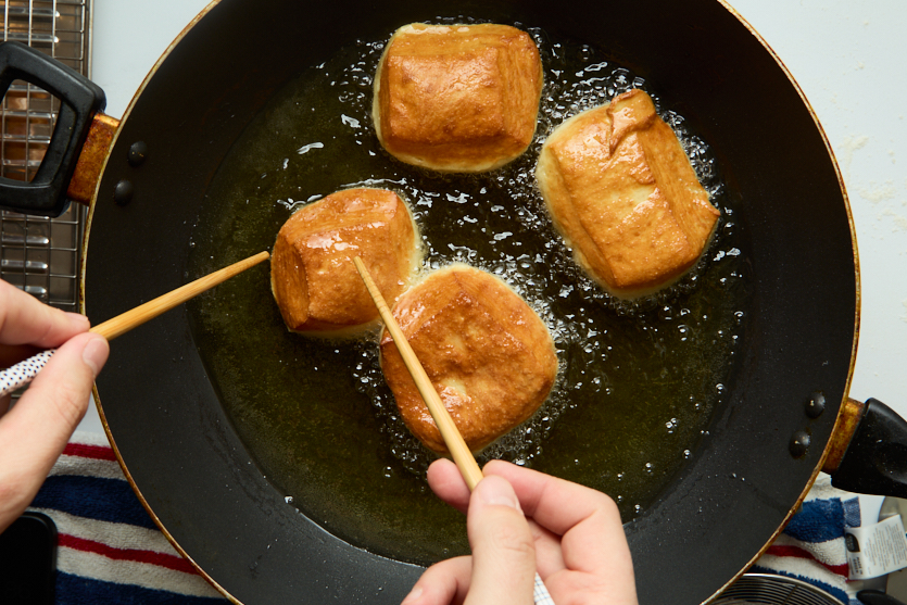 Four square pieces of dough, soon to become crispy beignets, fry in hot oil inside a black pan. A person uses two chopsticks to turn one of the pieces. A striped towel and wire rack are partially visible nearby.