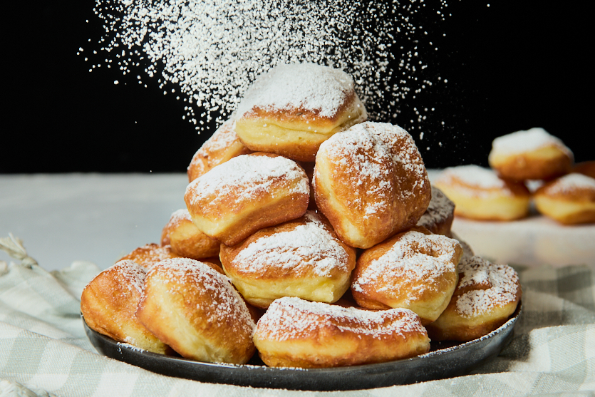 A plate of square-shaped New Orleans beignets stacked in a pyramid, dusted with powdered sugar falling from above. The chewy beignets rest on a checkered cloth, and more beignets are visible in the blurred background.