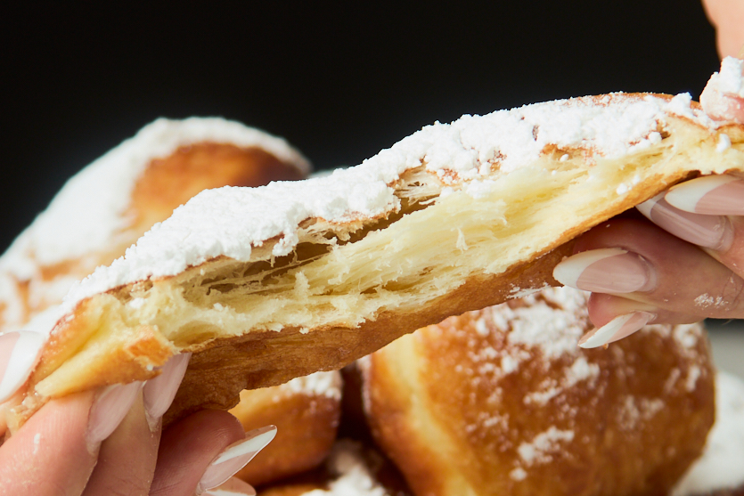 A close-up of hands pulling apart a flaky pastry covered in powdered sugar, revealing its airy layers—reminiscent of crispy beignets—while similar pastries blur softly in the background.