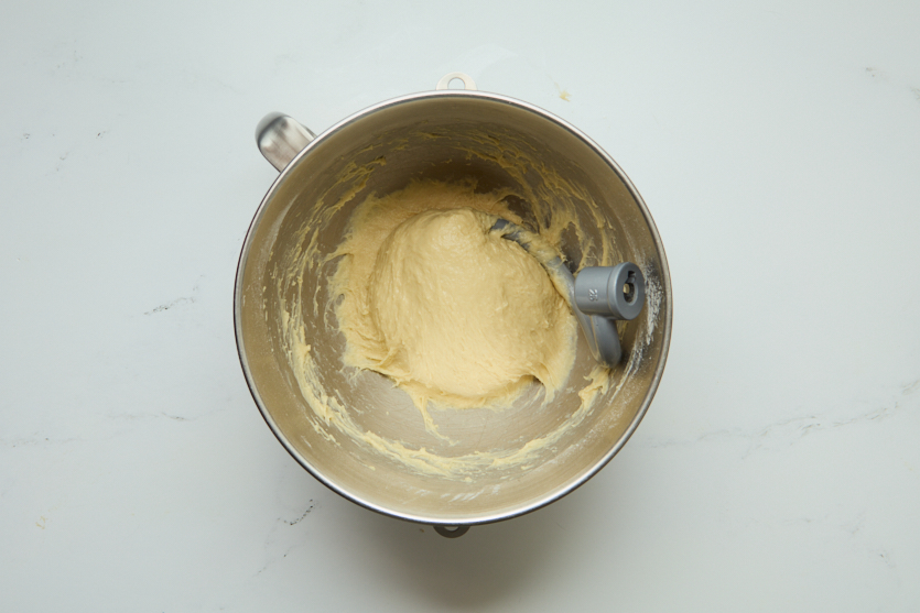 A mixing bowl with pale yellow dough for chewy beignets and a dough hook attachment on a white surface. Some dough is sticking to the sides of the bowl, and the mixture appears smooth and thick, ready to be transformed into classic New Orleans beignets.