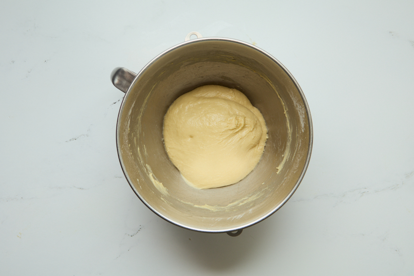 A metal mixing bowl sits on a white surface, containing a smooth, round ball of uncooked dough. The dough appears to have just finished mixing and is resting in the center of the bowl, ready to be transformed into chewy beignets.