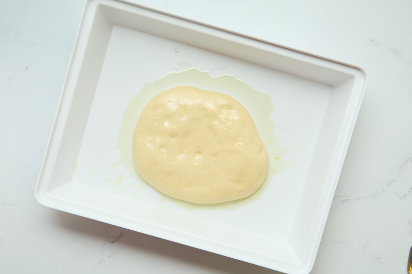 A rectangular white tray holds a round, slightly bubbly yellow dough spread in the center, surrounded by a thin layer of oil on a white surface—reminiscent of classic New Orleans beignets awaiting the perfect rise before transforming into irresistibly chewy beignets.