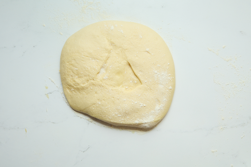 A piece of pale, slightly floured dough rests on a white surface. There is a faint triangle-shaped indentation in the center of the dough—hinting at the pillowy texture and signature look of New Orleans beignets before they’re fried to perfection.