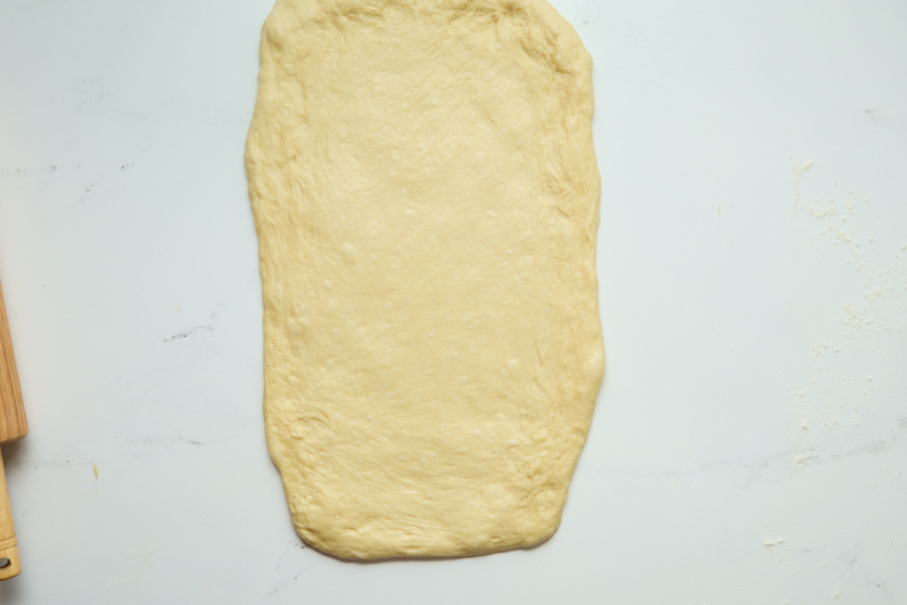 A sheet of uncooked dough is rolled out into a rough rectangle shape on a white surface, ready to be transformed into chewy beignets. The edges are uneven, and a wooden rolling pin is partially visible in the corner.