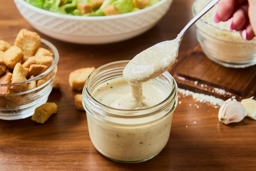 A spoon lifts creamy Caesar salad dressing from a glass jar. Surrounding the jar are bowls of croutons, shredded cheese, garlic cloves, and a salad in the background, all set on a wooden table.