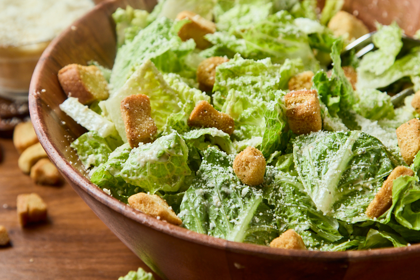 A wooden bowl filled with Caesar salad, featuring romaine lettuce, croutons, and grated cheese. Some croutons are scattered on the table next to the bowl.