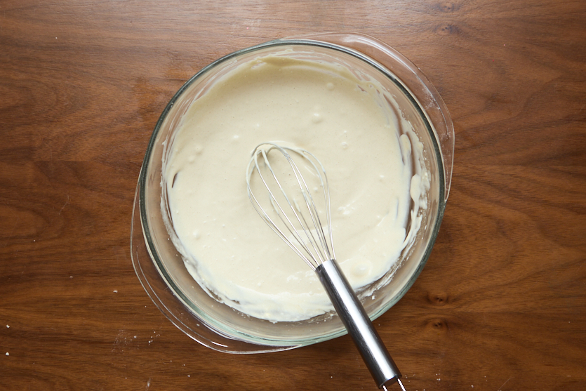 A glass mixing bowl filled with thick, pale batter sits on a wooden surface. A metal whisk rests inside the bowl with some batter clinging to it.