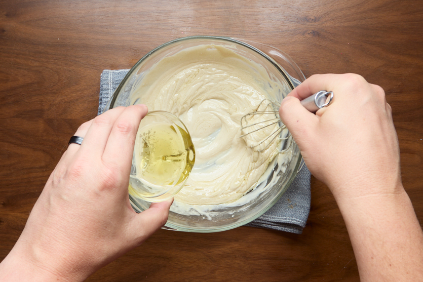 A person pours a clear liquid from a small glass bowl into a larger bowl of batter while whisking, on a wooden surface with a gray cloth underneath.