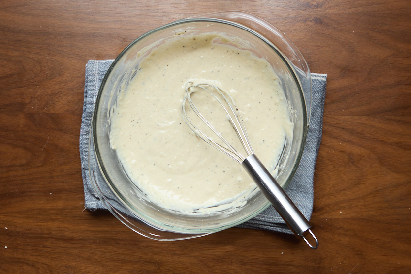 A glass bowl containing a creamy batter with visible specks and a metal whisk rests inside. The bowl sits on a folded blue cloth atop a wooden surface.