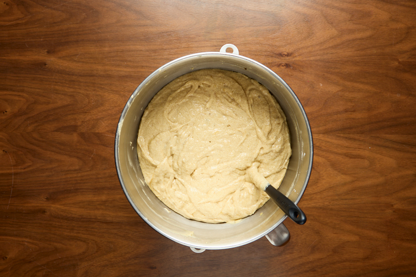 A metal mixing bowl filled with thick, light-colored batter sits on a wooden surface. A black-handled spatula rests inside the bowl.