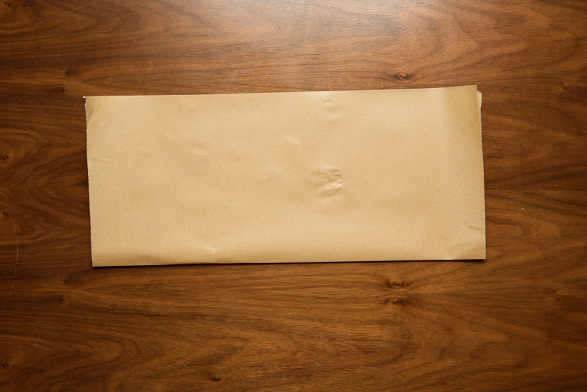 A rectangular sheet of brown paper is placed flat on a wooden surface. The paper has slight creases and appears unmarked. The wood has a visible grain with warm tones.