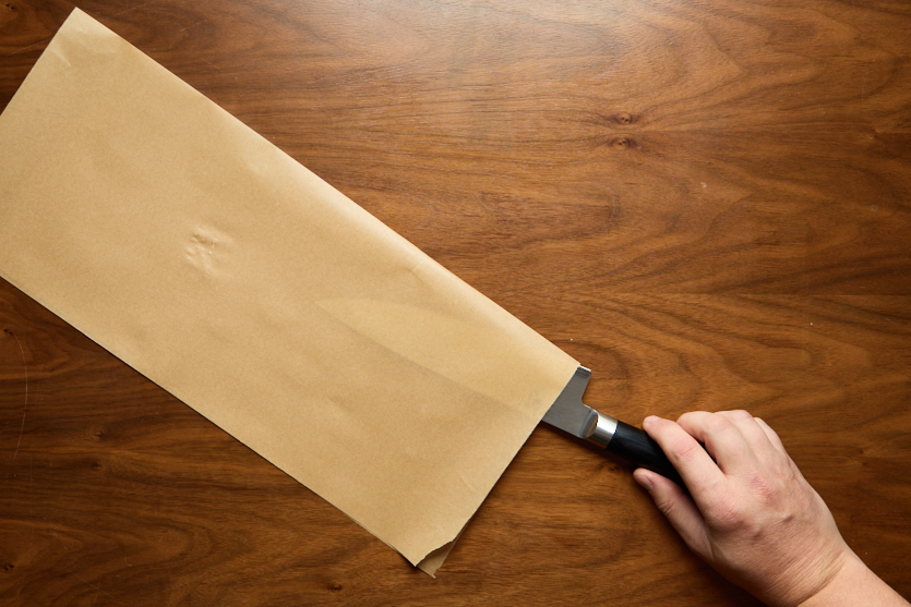 A hand holds a kitchen knife partially covered with a large sheet of brown parchment paper, placed on a wooden surface.