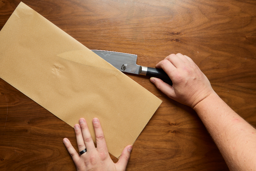 A person holds a chefs knife in one hand and a piece of brown parchment paper in the other, preparing to test the knifes sharpness on the paper. The background is a wooden surface.